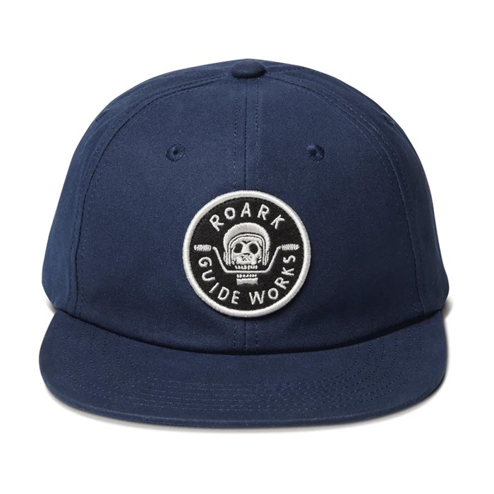 straight on view of a blue baseball cap woth a small circular patch in the center of the hat. The patch has a skull biting motorcycle handle bars with the word "roark" at the top of the circle and "guide Works" at the bottom.