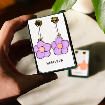 image of a hand holding a small box with a earring card displaying two earring. The earrings are light purple flowers with orange centers. 