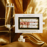 Small white box, sits atop a gold draped piece of silk. There are three champagne cubes displayed out side of the box with a tall glass fizzing next to it. 
