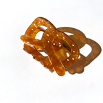 IMAGE SHOWS A HAIR CLIP IN THE SHAPE OF A KNOTTED PRETZEL. THERE ARE SMALL HAND PAINTED SALT FLAKES AND A BROWN SHADOW BEING CASTED BEHIND THE CLIP. BROWNISH AMBER COLORED CLIP AGAINST A WHITE BACKGROUND. 
