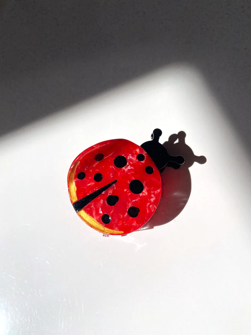 LADY BUG CLAW CLIP SITTING ON A WHITE BACKGROUND. RED CIRCLE WITH10 BLACK DOTS OF VARIOUS SIZES WITH A SMALL BLACK PIE SHAPE CUT OUT TO DEFINE THE LADYBUGS WINGS. 
