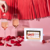 a pink backdrop with bright red tassels hanging from up top. A hand is dropping a mimosa cube into a glass of champagne,. There are rose petals, raspberries and pink sugarcubes on the floor. 