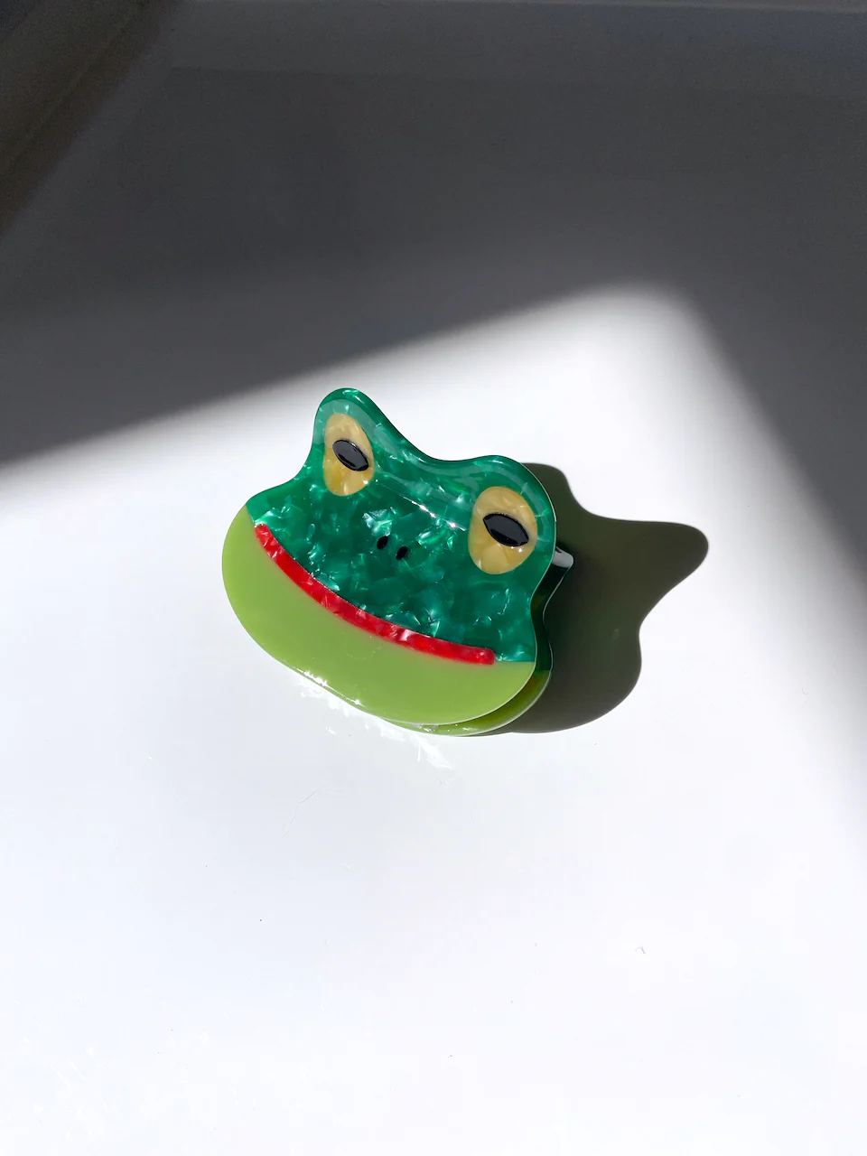 GREEN HAIR CLIP WITH DIFFERENT COLORED PARTS MADE TO RESEMBLE A SMILING FROG. IT HAS BROWN EYES AND A SMILING RED MOUTH. 