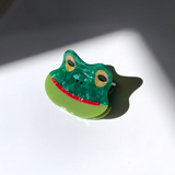GREEN HAIR CLIP WITH DIFFERENT COLORED PARTS MADE TO RESEMBLE A SMILING FROG. IT HAS BROWN EYES AND A SMILING RED MOUTH. 