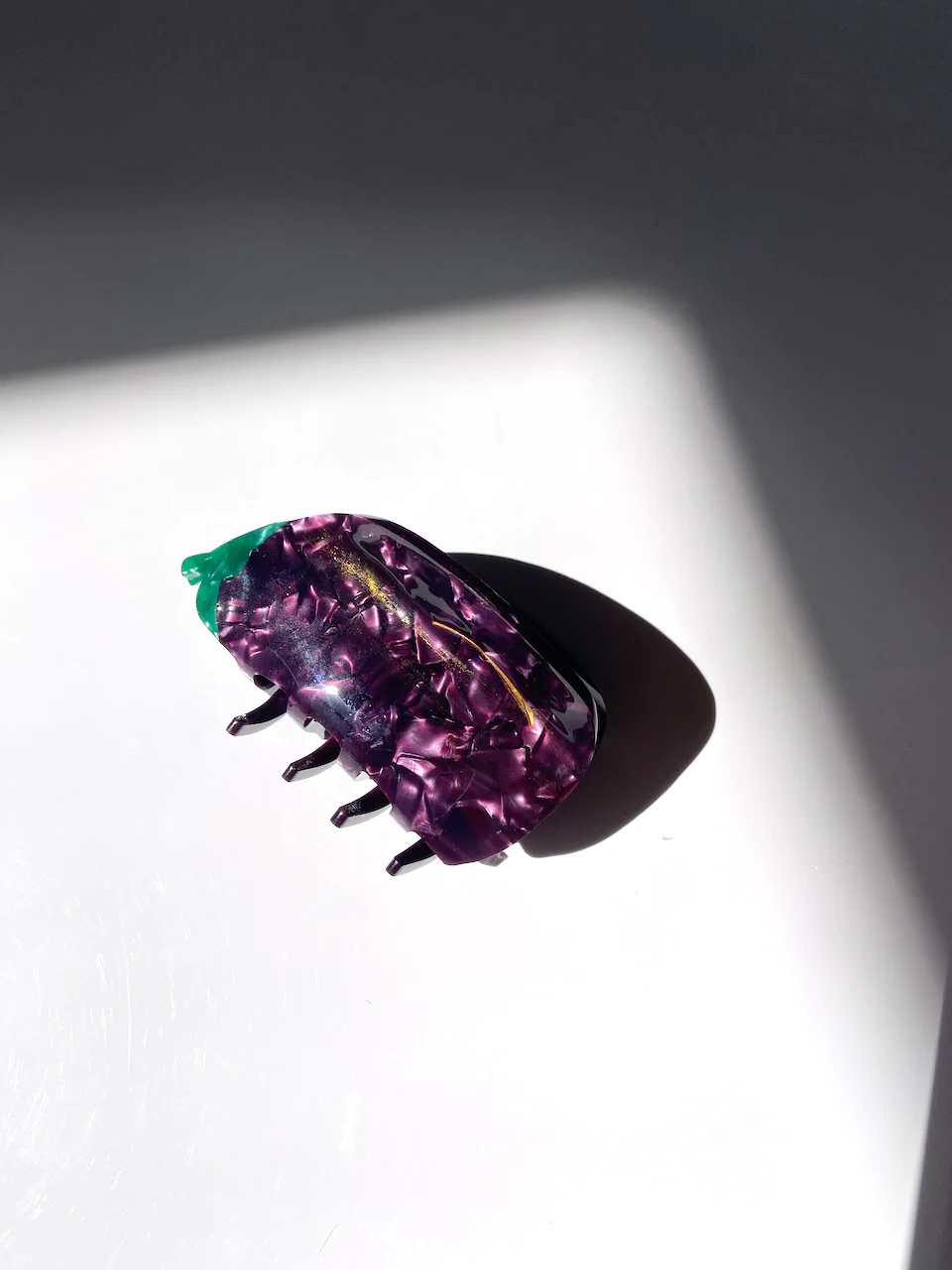 A MARBELED PURPLE CLAW CLIP WITH A GREEN TOP MADE TO LOOK LIKE AND EGGPLANT. THE CLIP SITS ON A WHITE BACKGROUND WHICH SHOWS THE DEPTH OF COLOR. 