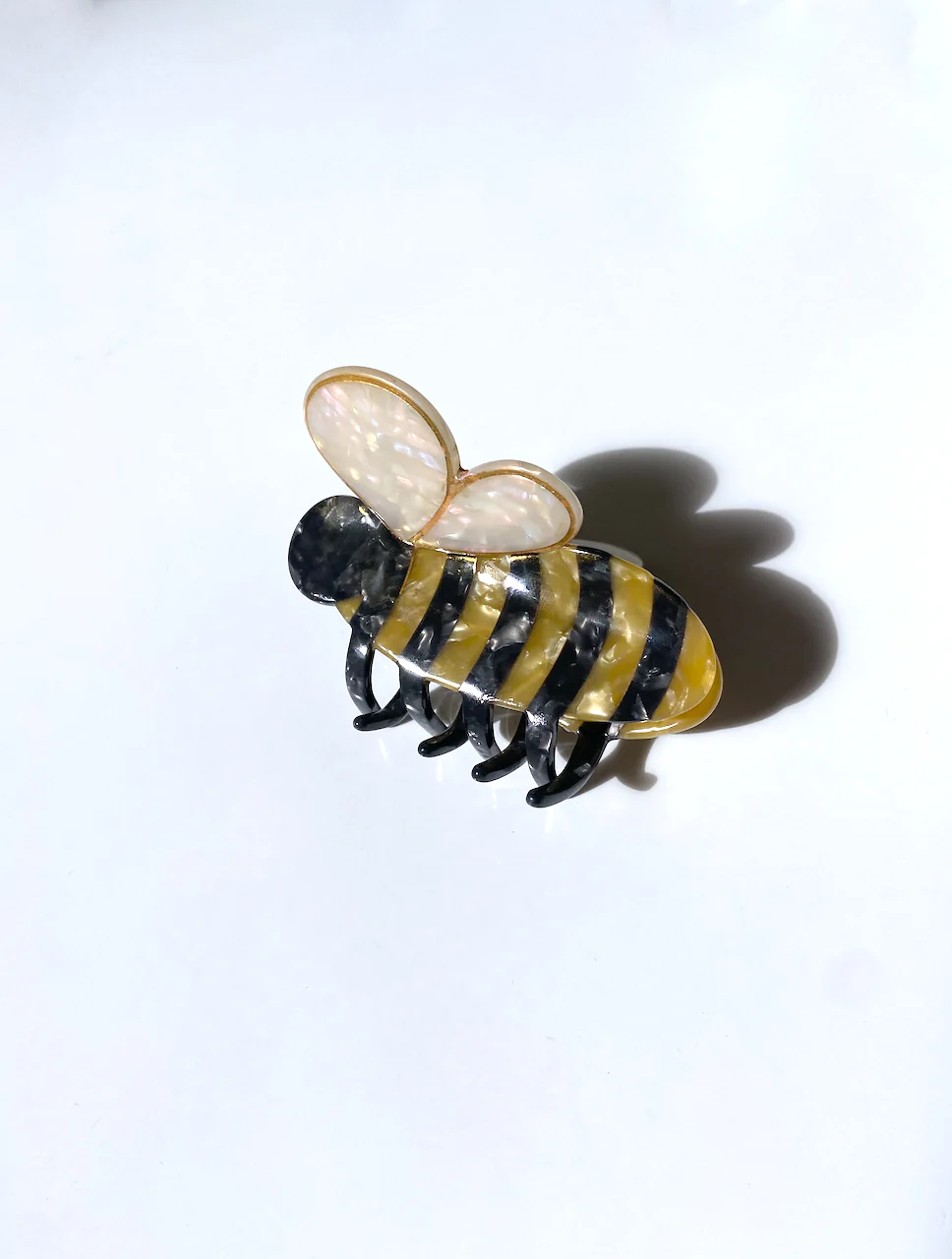 HAIR CLIP WITH A YELLOW AND BLACK STRIPED BODY, AND AN OFFWHITE HEART SHAPE THAT IS MADE TO RESEMBLE A BUMBLE BEE. THE COLORFUL CLIP SITS ON TOP OF A WHITE BACKGROUND. 