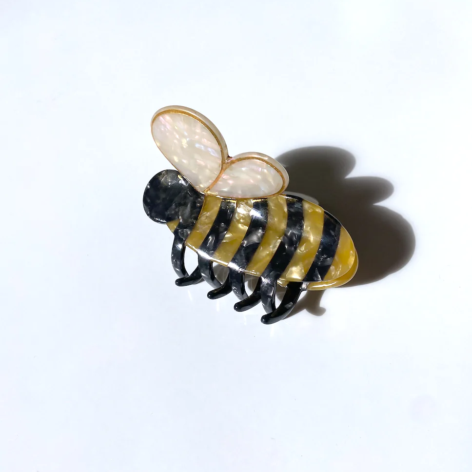 HAIR CLIP WITH A YELLOW AND BLACK STRIPED BODY, AND AN OFFWHITE HEART SHAPE THAT IS MADE TO RESEMBLE A BUMBLE BEE. THE COLORFUL CLIP SITS ON TOP OF A WHITE BACKGROUND. 