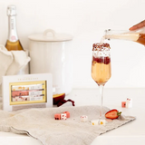 a white backdrop with a tan linen napkin laid out. A box of Teaspressa , sugar cubes and a mimosa sit on the napkin. The mimosa is being poured into. 