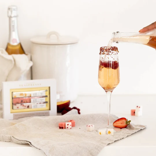 a white backdrop with a tan linen napkin laid out. A box of Teaspressa , sugar cubes and a mimosa sit on the napkin. The mimosa is being poured into. 