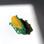 A YELLOW CORN HAIR CLAW CLIP WITH GREEN HUSKS AND GOLD DETAILING SITS IN FRONT OF A WHITE BACKGROUND. YOU CAN SEE DETAILING IN THE HUSK LEAVES AS WELL AS THE CORN KERNELS. 