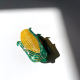 A YELLOW CORN HAIR CLAW CLIP WITH GREEN HUSKS AND GOLD DETAILING SITS IN FRONT OF A WHITE BACKGROUND. YOU CAN SEE DETAILING IN THE HUSK LEAVES AS WELL AS THE CORN KERNELS. 