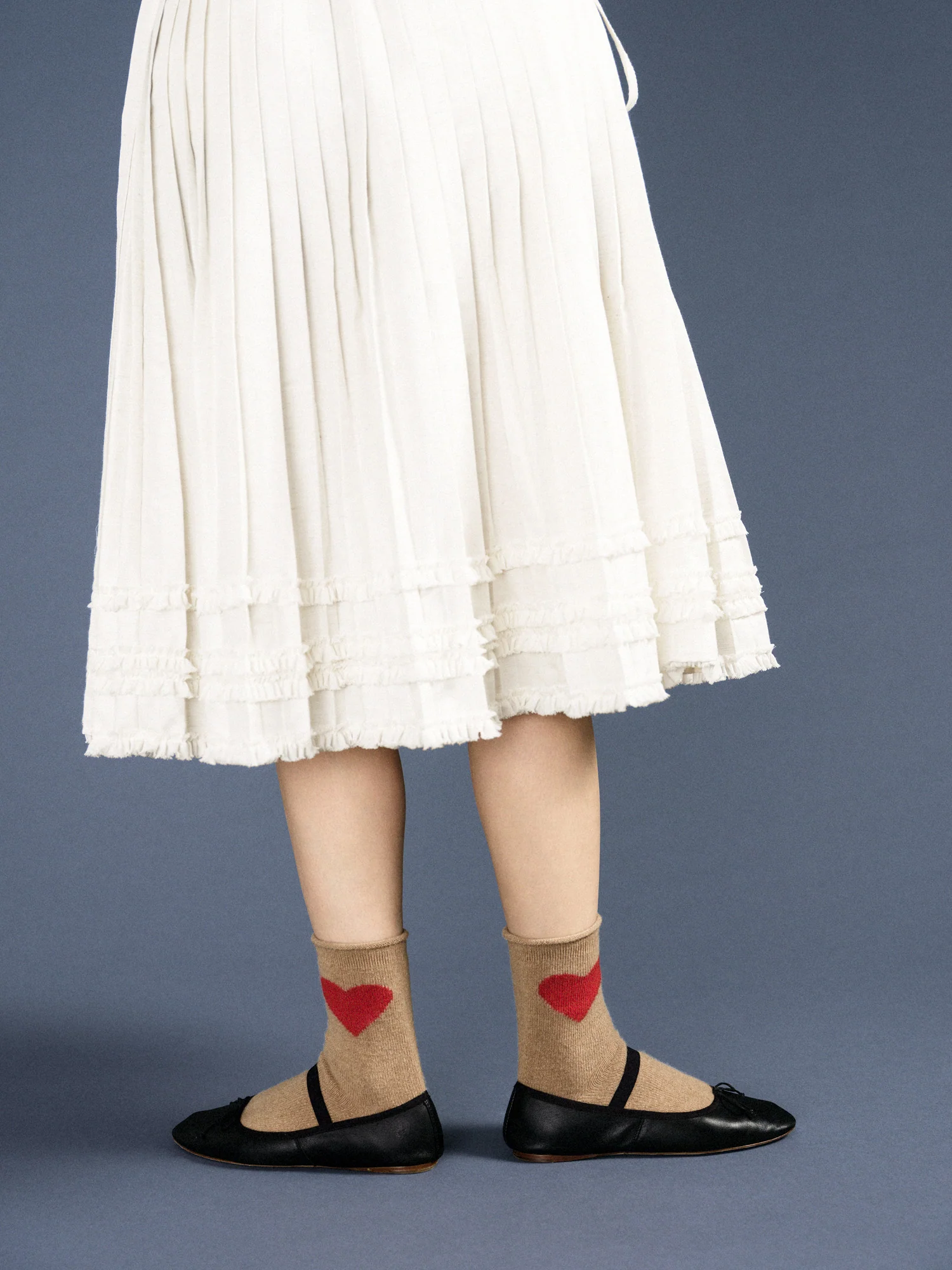 a person wearing a white pleated skirt. The camel colored sock is paired with a black ballet flat. The big bright red heart pops against some of the other colors in the picture. 