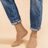 person wearing blue jeans and tan socks stands in front of a light colored background. 