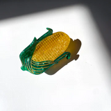 A YELLOW CORN HAIR CLAW CLIP WITH GREEN HUSKS AND GOLD DETAILING SITS IN FRONT OF A WHITE BACKGROUND.